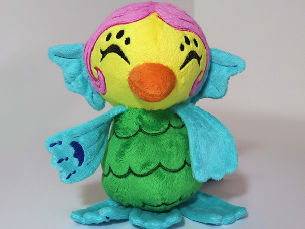 A photo of an amabie plushie. Its face is yellow, with a orange beak, pink hair styled into a bun, and blue ear fins. Its body is green with a scales pattern, and it has 2 blue arm fins, and 3 blue leg fins.