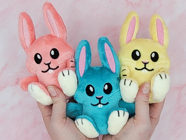 two hands holding three small bunny plushies. They are pink, blue, and yellow and the size of the person's palm.