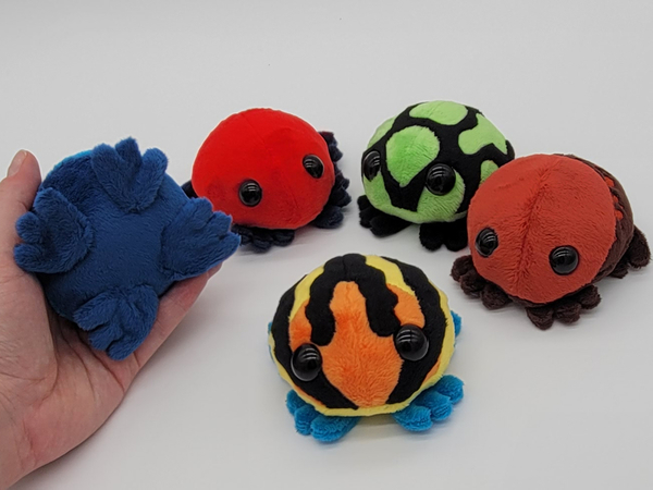 Five frog plushies sitting in a semi circle facing the viewer. They are brightly colored to look like amazonian poison dart frogs. A hand is holding one up to the camera to show off the feet on the underside.