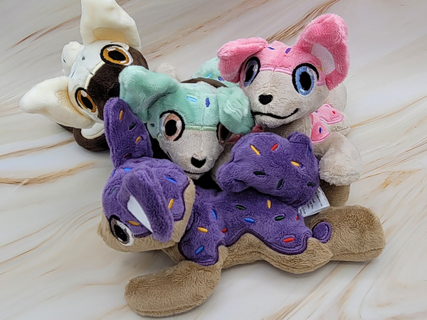 A group of dog plushies that have frosting and sprinkles covering their backs. There is a dark tan dog with purple frosting; a chocolate brown dog with white frosting; and two light tan dogs. One has pink frosting and the other has mint green frosting.