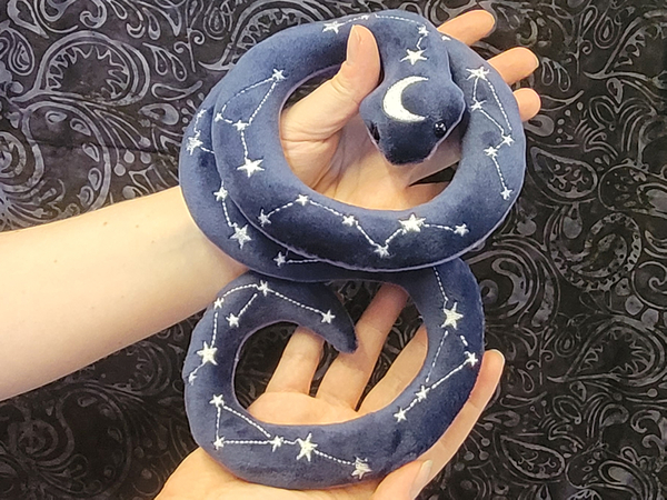 Two hands holding up a snake plushie. It is blue and has stars on the back arranged like constellations.