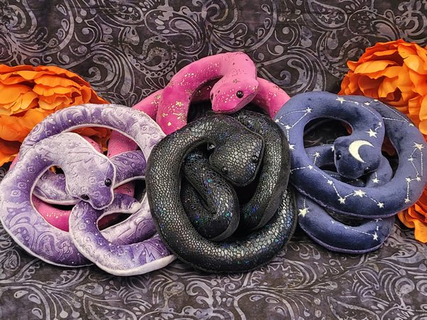 A pile of four snake plushies. There is a magenta snake with gold flecks, a black snake with iridescent scales, a light purpe snake with small gems, and a blue snake with stars on it.